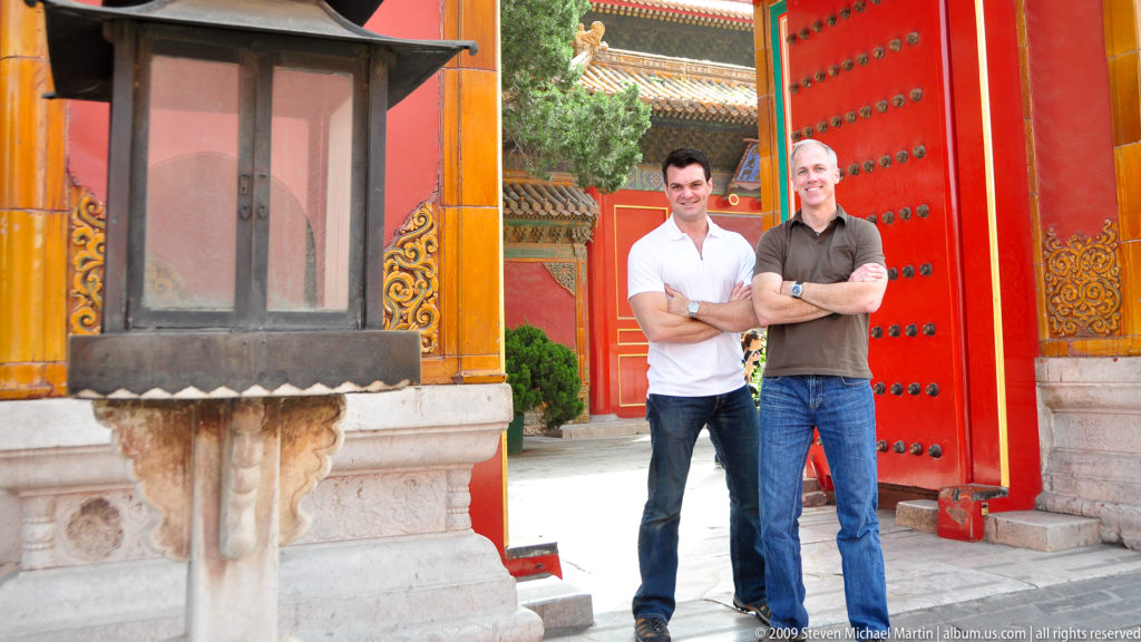Two male visitors stand with arms crossed in a red, yellow-trimmed doorway in the Forbidden City.