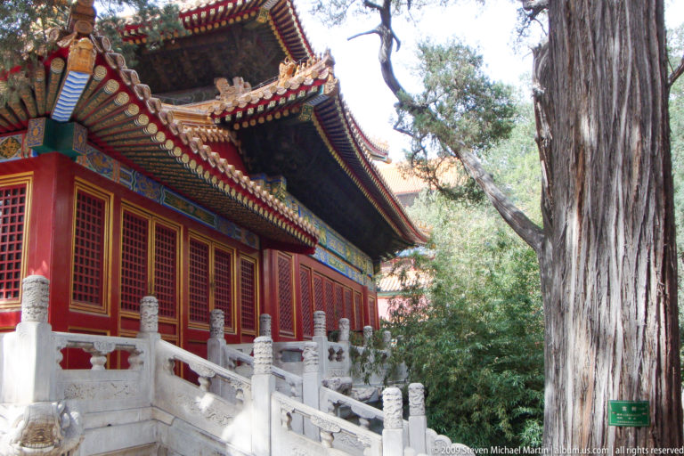 Side view of a red timber hall with lattice windows and painted eaves above a white stone balustrade, partly obscured by mature trees in a palace garden.