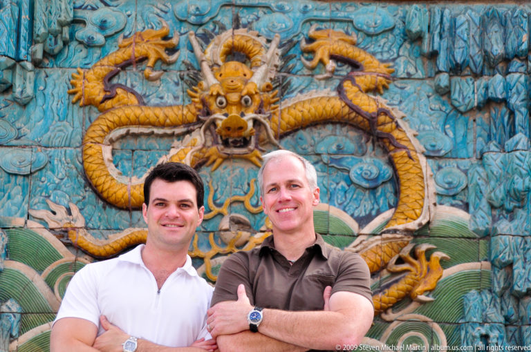 Two male visitors smiling in front of a large yellow dragon relief on the Forbidden City’s Nine-Dragon Screen.