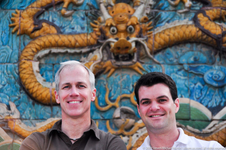 Two male visitors smiling in front of a yellow dragon relief on the Forbidden City’s Nine-Dragon Screen.