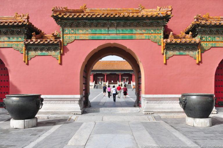 Arched gateway in a red palace wall with large bronze water vats on stone bases on either side.