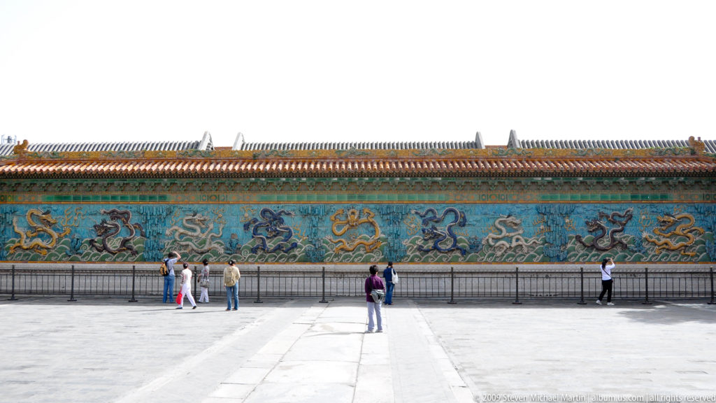 Wide frontal view of the Forbidden City Nine-Dragon Screen, a long turquoise glazed wall with nine relief dragons beneath a yellow-tiled roof on a stone base.