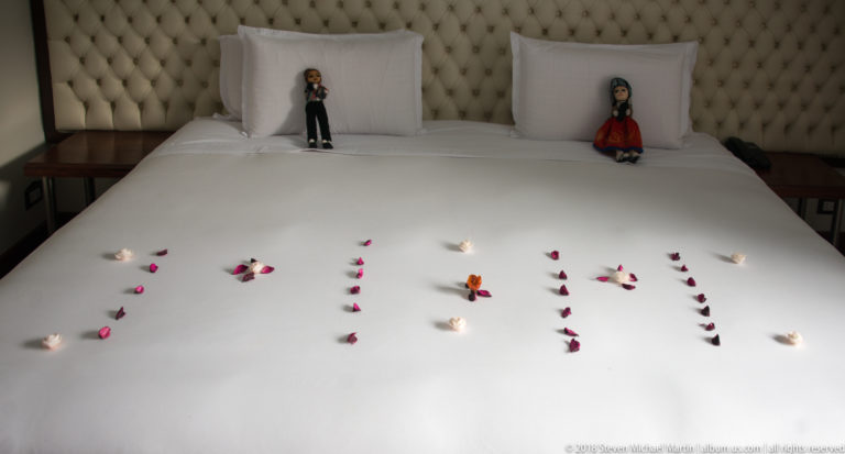 White hotel bed decorated with flower petals, with two handmade textile dolls placed upright against the pillows.