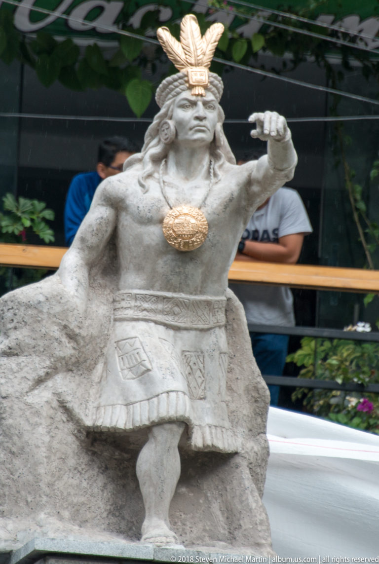 Seated stone figure in Inca-style regalia with feathered headdress, patterned tunic, cape, and large chest ornament.