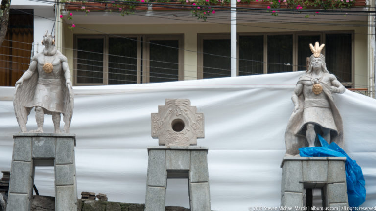 Two Inca-themed stone figures on pedestals flank a carved geometric stone element in an outdoor public setting.