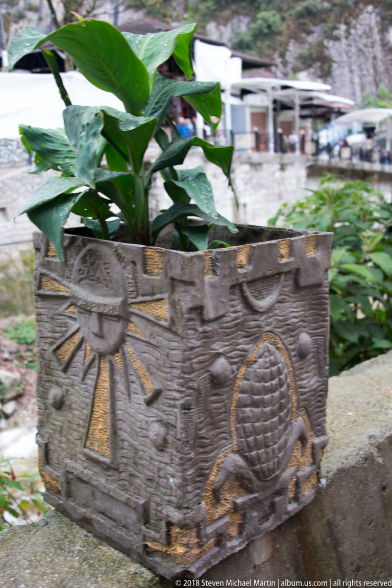 Rectangular planter with raised sun, geometric, and maize motifs, holding broad green plants.