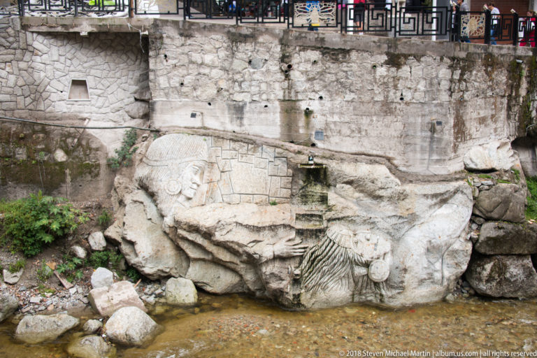 Large carved stone head and shallow relief beside a stream, with water issuing from openings in a retaining wall.