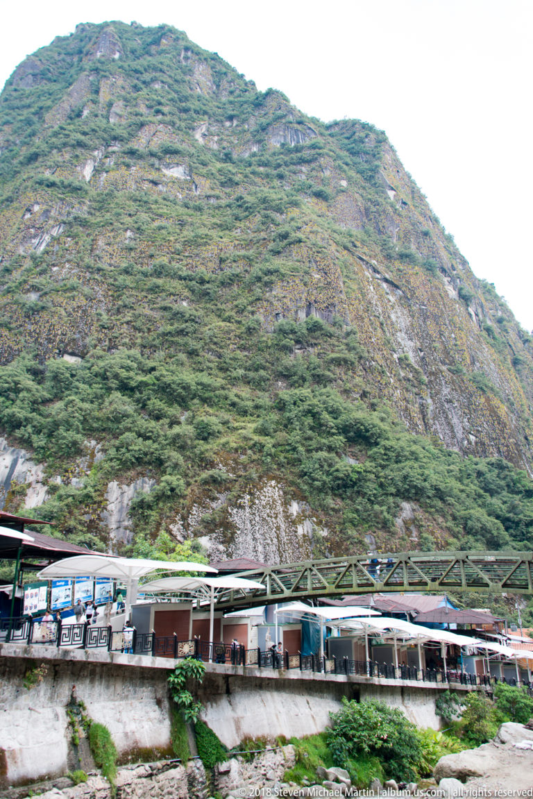 Passenger train and low railway structures beneath a massive steep mountain covered in dense green vegetation.