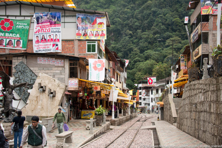 Railway tracks running between shops, retaining walls, benches, and campaign posters in central Aguas Calientes.