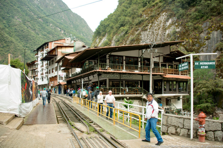 Rail tracks and pedestrian path with yellow railings passing restaurants beneath steep mountain walls in Aguas Calientes.