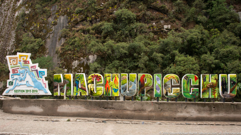 Large colorful letters spelling MACHUPICCHU beside a municipal emblem against a forested rock wall.