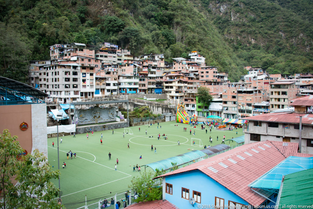 Elevated view of a green sports field surrounded by dense hillside buildings and steep forested mountains in Aguas Calientes.