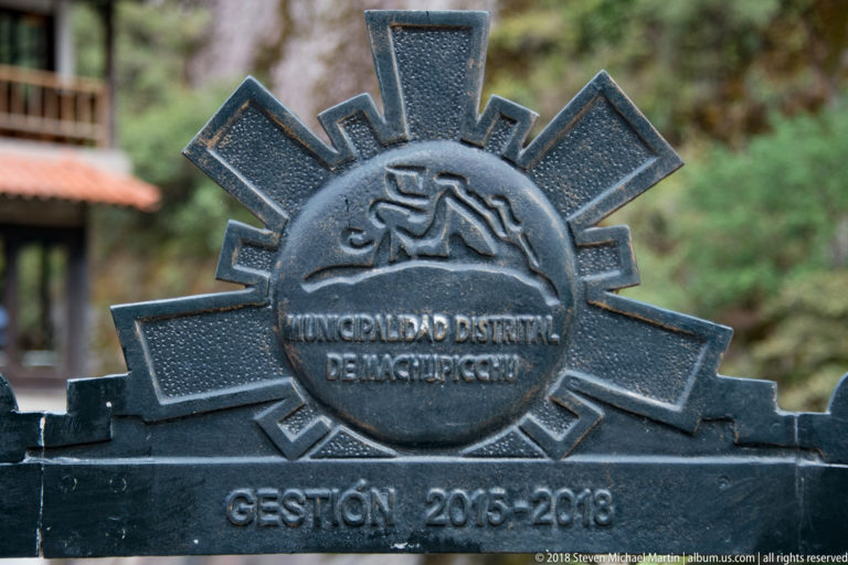 Dark metal emblem reading Municipalidad Distrital de Machupicchu and Gestión 2015–2018, with geometric rays.