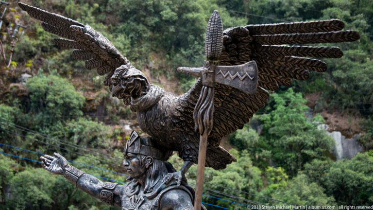 Bronze warrior-like figure holding a staff beneath a large bird with outstretched wings.