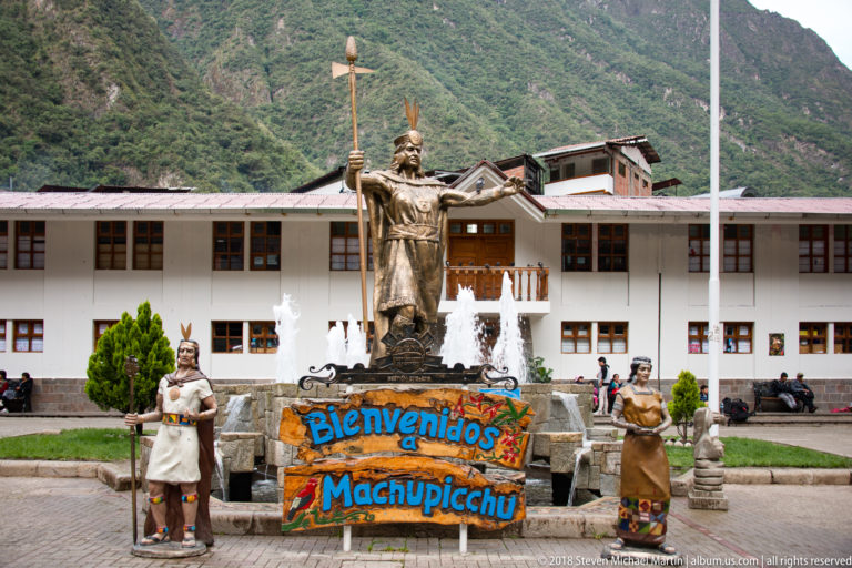 Bronze Inca ruler and attendants above a fountain with a colorful “Bienvenidos a Machupicchu” sign.