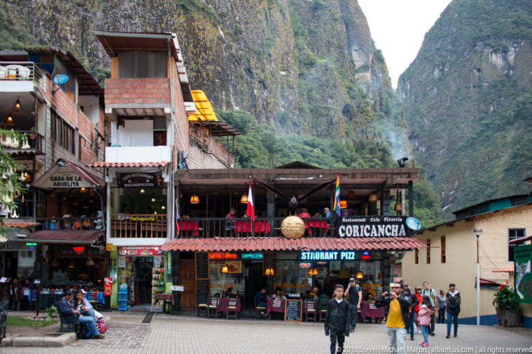 Coricancha restaurant and neighboring storefronts facing a pedestrian square beneath steep forested cliffs.