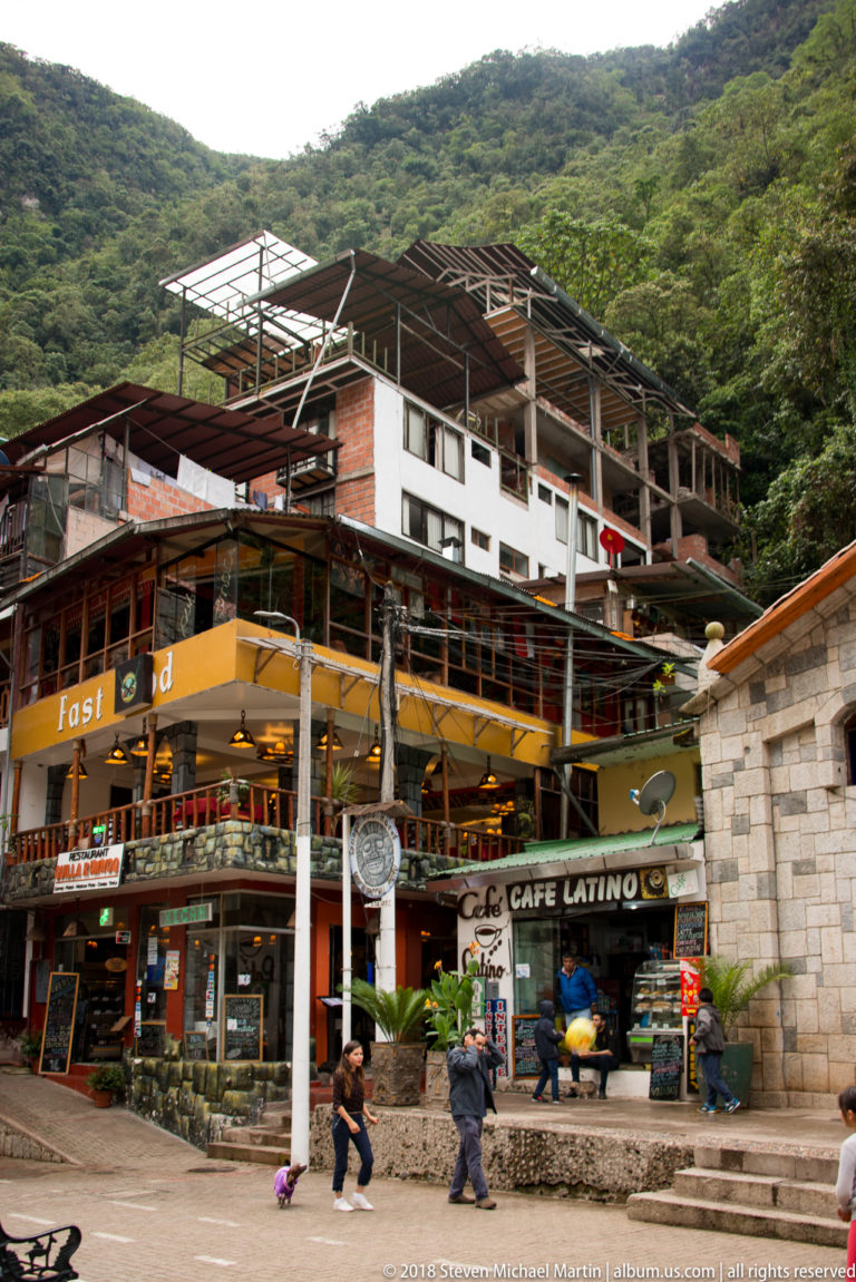Multistory cafés and restaurants beneath a steep forested hillside, with open terraces and unfinished roof framing.