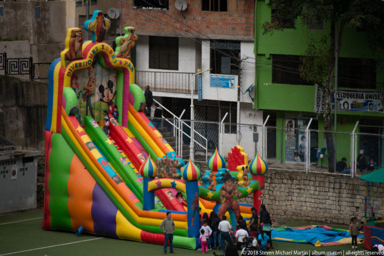 Large multicolored inflatable children's slide on a sports field with families gathered at its base and buildings behind.
