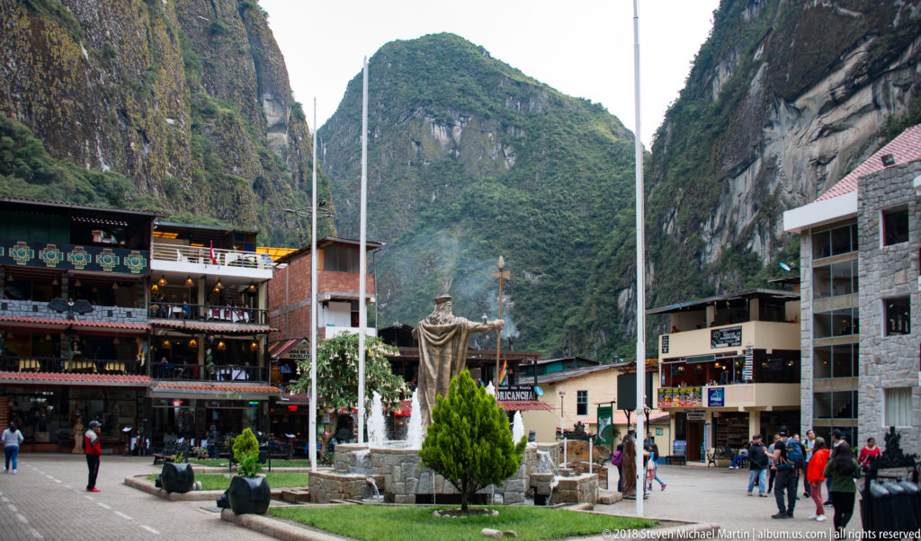 Bronze Inca ruler monument with fountains and flagpoles in a plaza beneath steep green mountains.