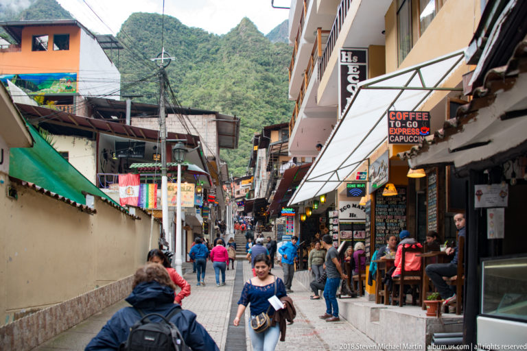 Crowded pedestrian street lined with cafés and shops, with a steep forested mountain rising at the far end.