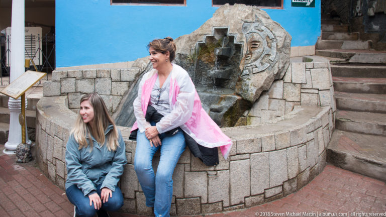 Two women seated beside a stone fountain with carved animal head, stepped water channel, and sun motif.