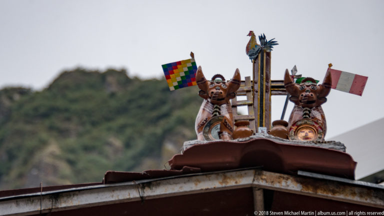 Two horned rooftop figurines holding Peruvian and multicolored Andean flags against a green mountain.