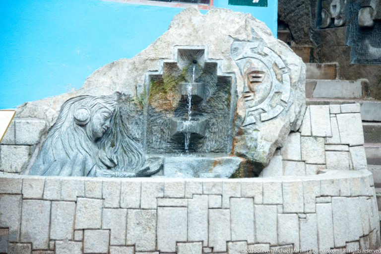 Stone fountain with carved lion-like head, stepped water channel, circular sun motif, and block masonry basin.