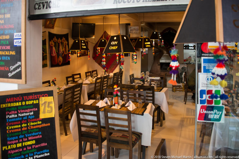 Restaurant interior with wooden tables, black pendant lamps, colorful decorations, and a Spanish tourist-menu board.