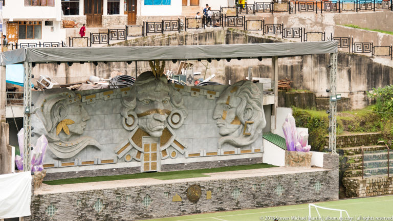 Large gray stage relief with a central mask-like face, two flanking profiles, and gold-colored geometric accents.