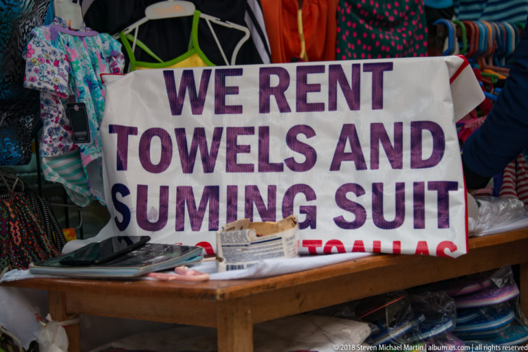 Hand-painted towel and swimsuit rental sign hanging before colorful clothing at a market stall.