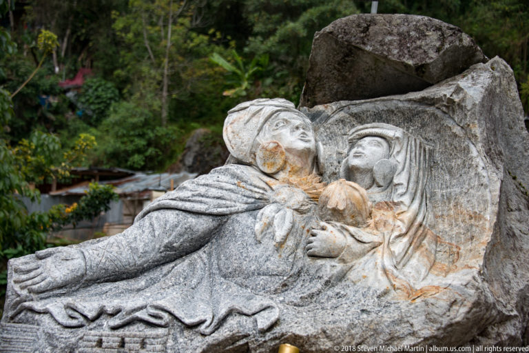 Carved stone group with a human figure and a larger face emerging from a partly rough boulder.