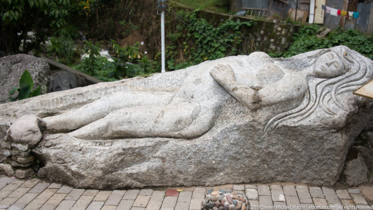 Large stone sculpture of a reclining nude woman carved along a horizontal boulder beside a paved path.