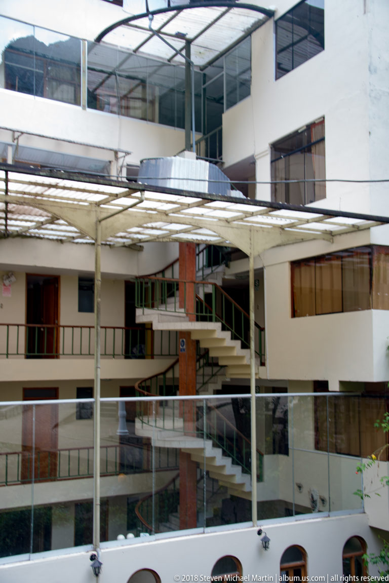 Modern white multistory courtyard building with glass railings, brick accents, metal canopy, and exterior stairways.