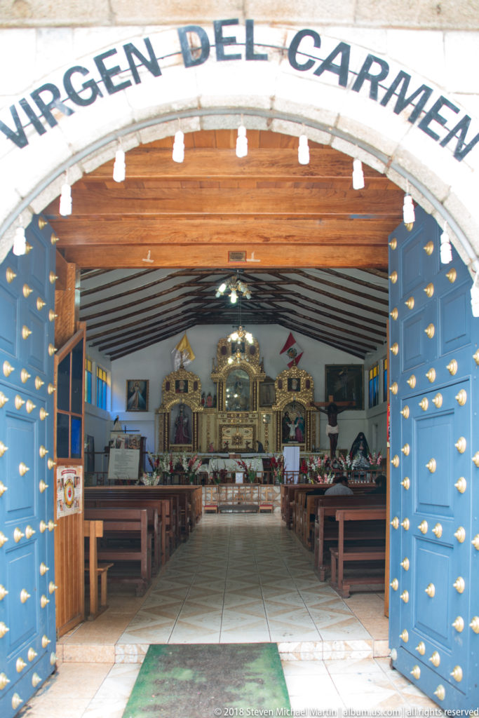 Open blue studded chapel doors framing wooden pews and a gilded altar beneath an entrance inscription ending “DEL CARMEN.”