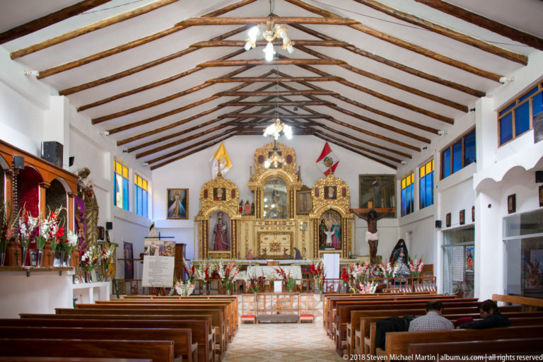 Long church nave with wooden pews, exposed timber rafters, blue windows, and a gilded altar at the far end.