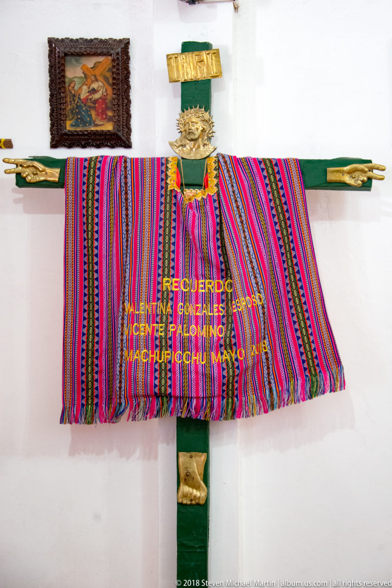 Wooden cross draped with a bright multicolored Andean textile and small gold-colored ornaments on a white wall.