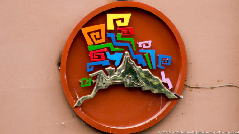 Circular ocher wall emblem with colorful stepped geometric motifs surrounding a raised green-and-gray mountain form.