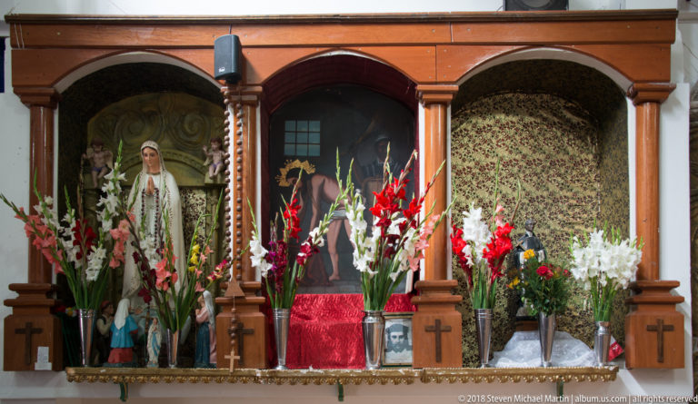 Wooden church shrine with a female devotional statue, flowers, patterned textile, and small framed religious images.
