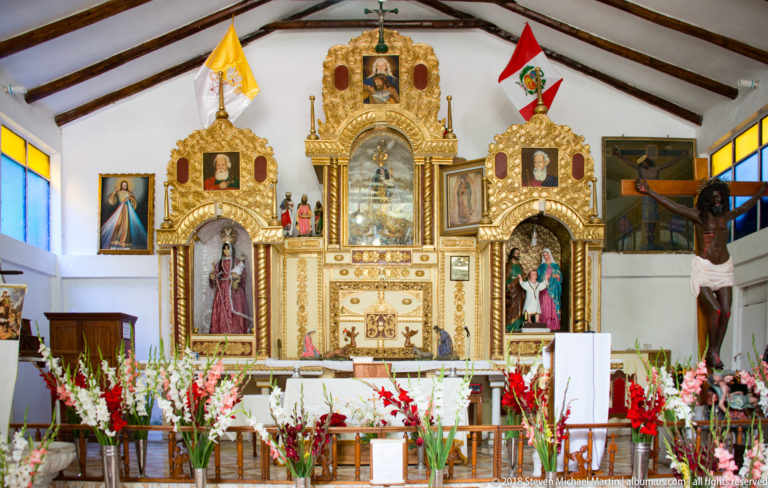 Wide view of a church sanctuary with gilded altar, crucifix, devotional images, timber roof, and abundant red and white flowers.