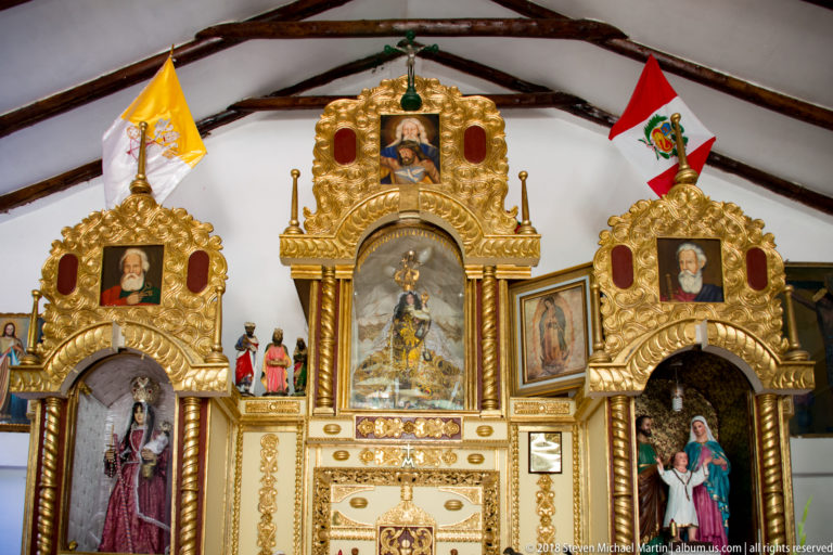 Ornate three-part gilded church retable with statues, devotional paintings, flags, and a central tabernacle-like structure.