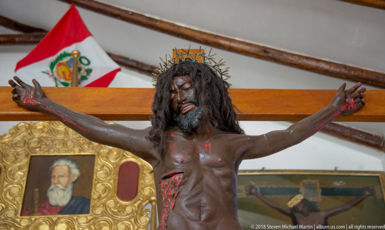 Polychrome crucifix of Christ wearing a crown of thorns and white loincloth, with gilded church furnishings behind.