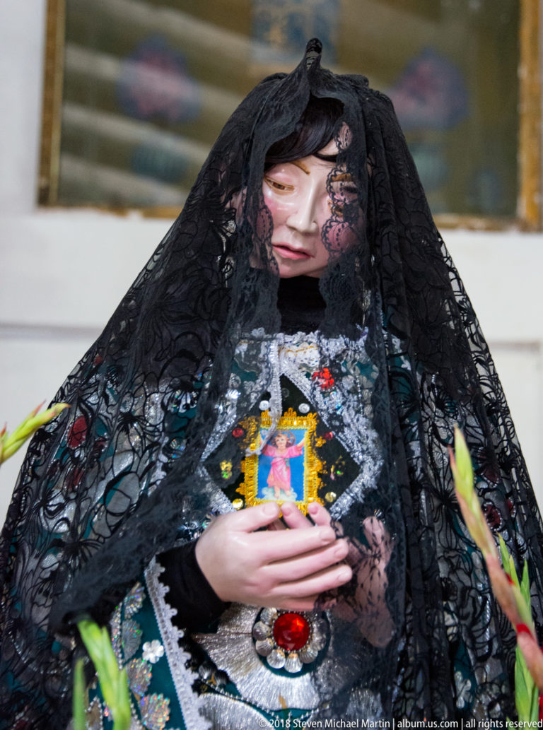 Veiled statue of the Virgin Mary in black lace and embroidered clothing, holding a small devotional image.