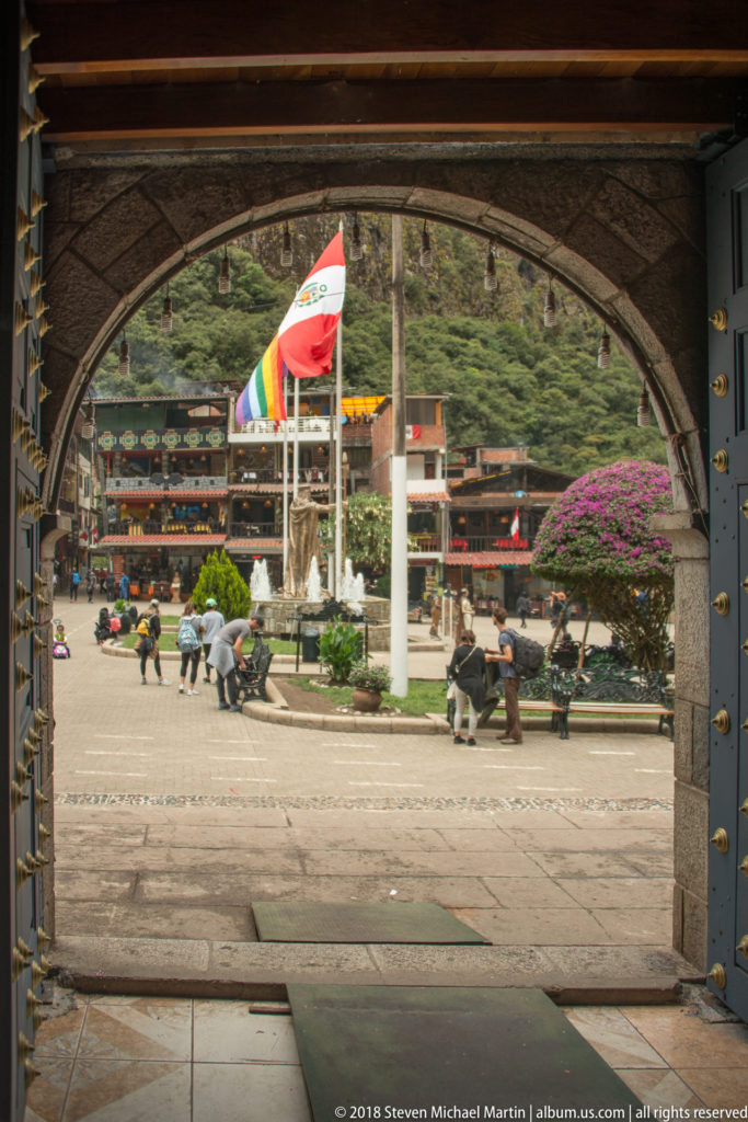 Stone archway framing a bright civic plaza with benches, flagpoles, people, flowers, and green mountains beyond.