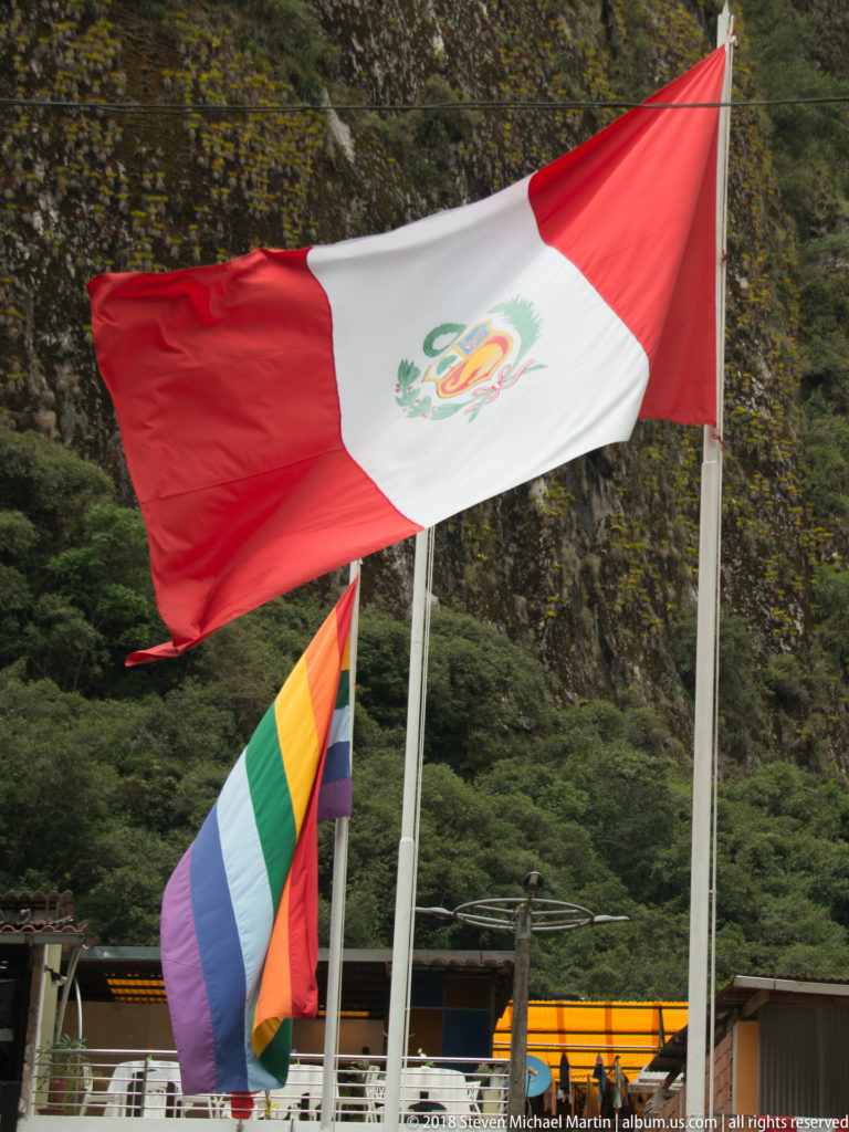 Peruvian red-and-white flag flying above a multicolored Andean flag against a steep green mountain.