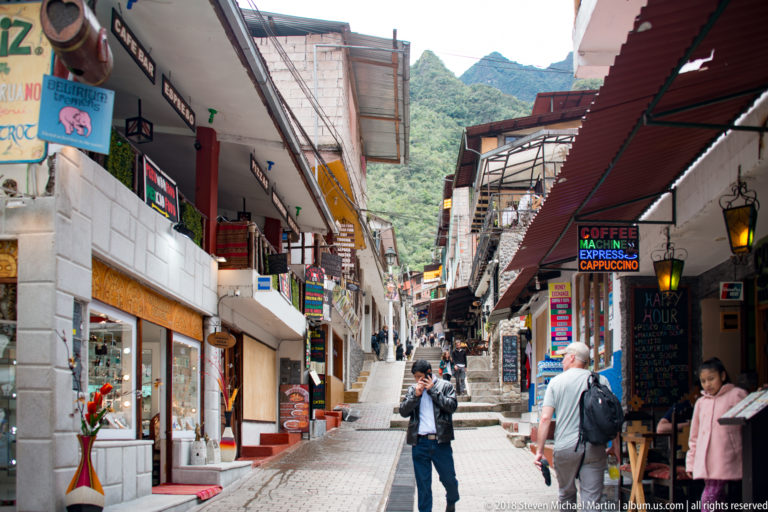 Pedestrians walking along a steep shop-lined lane with colorful signs and awnings in central Aguas Calientes.