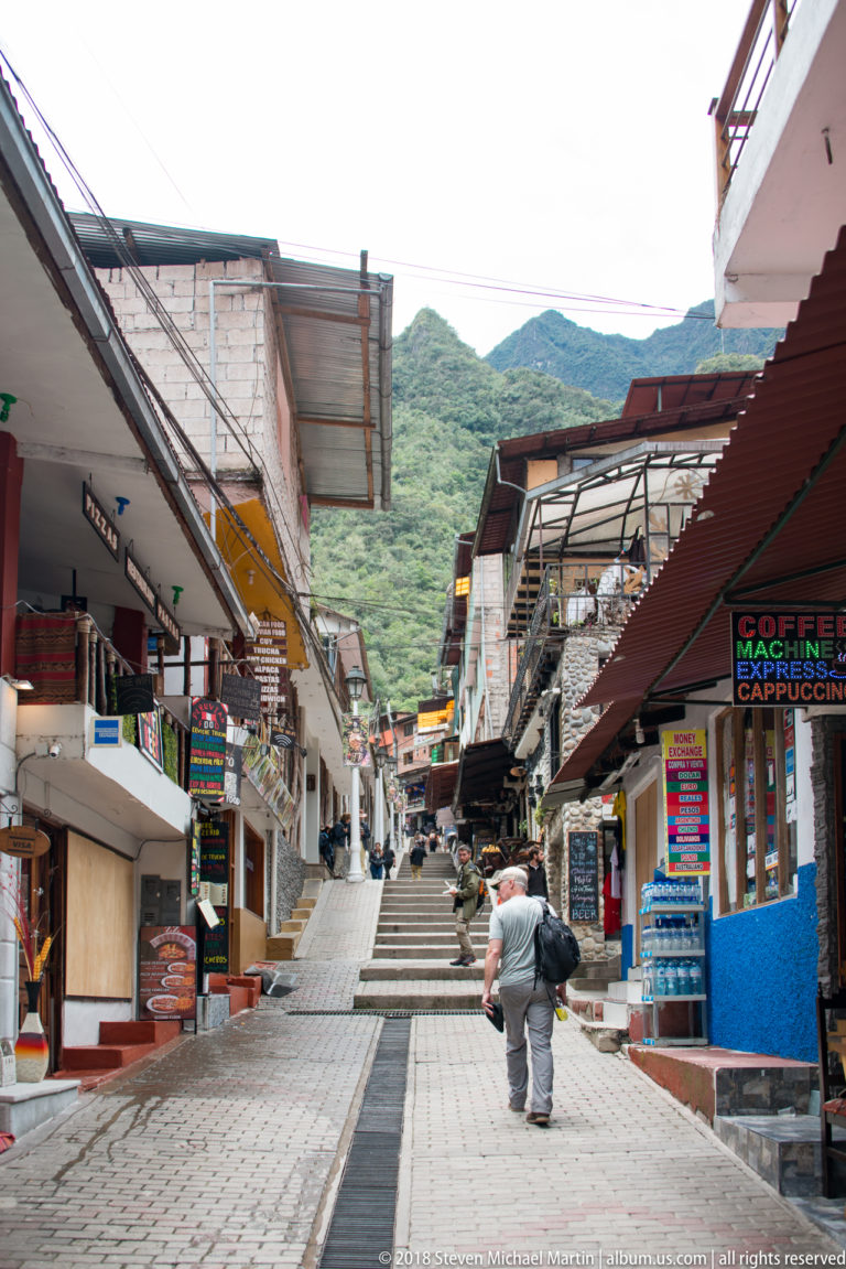 Steep commercial pedestrian street with shops, awnings, café signs, pedestrians, and a green mountain ridge beyond.