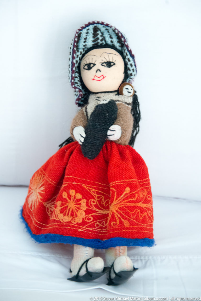 Handmade female textile doll in a red embroidered skirt and knitted head covering, carrying a small child.