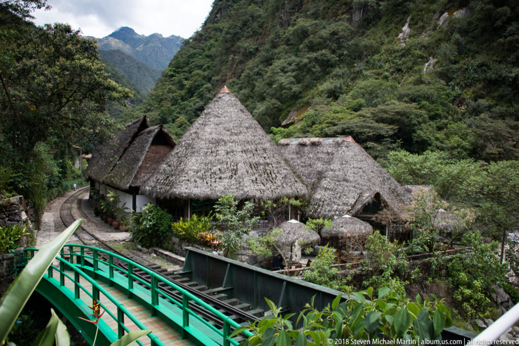 Thatched-roof buildings beside railway tracks and a green metal structure, surrounded by steep forested mountains.