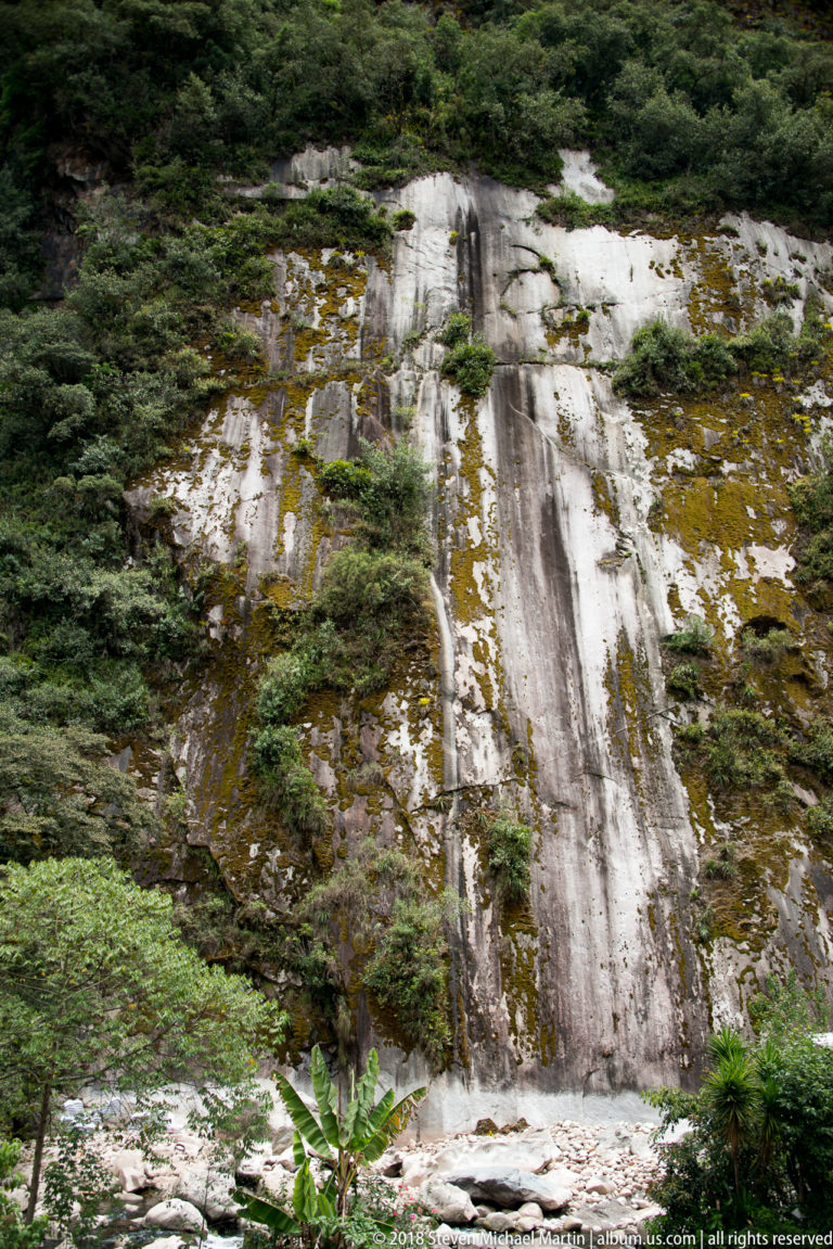 Thin waterfall flowing down a broad pale rock face bordered by dense green vegetation.