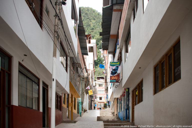 Steep narrow pedestrian lane between white and painted buildings, with shop signs and green mountains visible ahead.